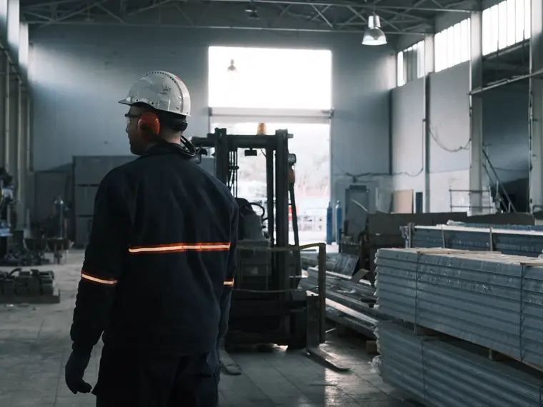 Ouvrier casqué dans une halle industrielle, chariot élévateur en fond.